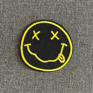 Puede incluir: Un parche circular negro con un borde amarillo que presenta un diseño de carita sonriente. La carita sonriente tiene los ojos tachados y la lengua fuera. El parche está sobre una superficie gris texturizada.