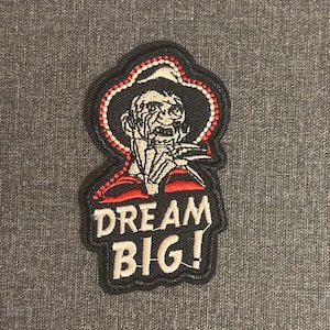 Peut inclure: Écusson brodé avec un bord noir, des accents rouges et blancs, et le texte "DREAM BIG!". L'écusson présente une figure stylisée avec un chapeau. La figure est en noir, rouge et blanc.