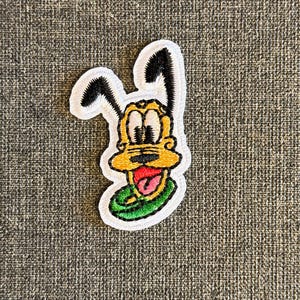 Op de afbeelding: Geborduurde patch met een cartoonhond met gele vacht, zwarte oren en een rode tong. De patch heeft een witte rand en een groene kraag. Het ontwerp is een klassiek stripfiguur.