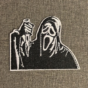 Peut inclure: Un écusson brodé en noir et blanc représentant le masque emblématique de Ghostface de la série de films d'horreur Scream. L'écusson représente la figure tenant un couteau, avec des coutures détaillées et une bordure blanche.