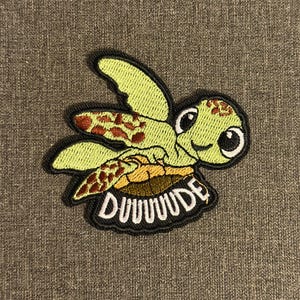 Puede incluir: Parche bordado con una tortuga de dibujos animados con una cara sonriente. La tortuga es de color verde claro con detalles marrones y naranjas. El parche tiene la palabra "DUUUUDE" en letras blancas sobre un borde negro. El parche está sobre un fondo gris.