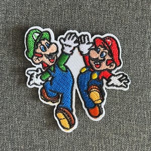May include: Embroidered patch featuring the characters Mario and Luigi from the Super Mario Bros. video game. Mario is wearing a red hat and Luigi is wearing a green hat. Both characters are giving a high five. The patch has a white border.