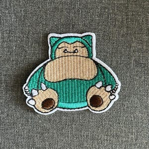 以下が含まれることがあります： 漫画のキャラクター、カビゴンの刺繍パッチ。パッチは緑色の体、ベージュのお腹、茶色の足で、白で縁取られています。パッチは、布製のアイテムにアイロンをかけたり、縫い付けたりするように設計されています。