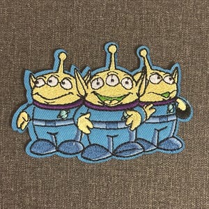 May include: Embroidered patch depicting three cartoon aliens with three eyes each. The aliens are wearing blue jumpsuits with a silver emblem. The patch has a blue border on a brown background.