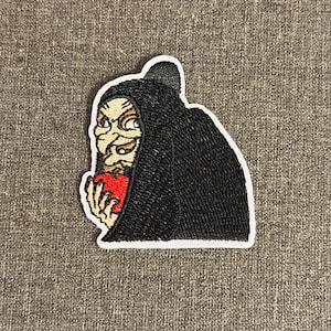 May include: Embroidered patch featuring a cartoon depiction of a witch in a black hooded cloak, holding a red apple. The witch has a sinister expression, with a white border around the patch. The patch is on a gray fabric background.