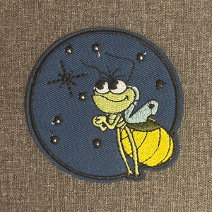 Może przedstawiać: Haftowana naszywka przedstawiająca kreskówkową świetlik z żółtym brzuchem, zieloną twarzą i niebieskimi skrzydłami. Świetlik jest umieszczony na ciemnoniebieskim okręgu z gwiazdą i małymi białymi kropkami, przypominającymi gwiazdy.