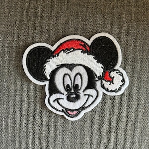 May include: Embroidered patch of Mickey Mouse wearing a Santa hat. The patch has a white border, black ears, and a smiling face. The Santa hat is red with a white trim. Ideal for decorating clothing or accessories.