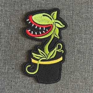 以下が含まれることがあります： 赤い裏地と白い歯を持つ大きな口を持つ漫画の植物を描いた刺繍パッチ。植物は黄色のアクセントが付いた緑色で、黄色のストライプが付いた黒いポットに入っています。パッチは灰色の背景にあります。