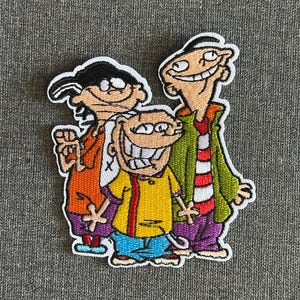 Puede incluir: Parche bordado con tres personajes de dibujos animados de la serie Ed, Edd y Eddy. Los personajes están delineados en negro con colores vibrantes, incluyendo naranja, amarillo, verde, morado y azul. El parche tiene un borde blanco.