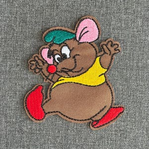 Puede incluir: Parche bordado con un ratón de dibujos animados en pose de carrera. El ratón es marrón con una camisa amarilla, zapatos rojos, un sombrero verde y orejas rosas. Fondo de tela gris.