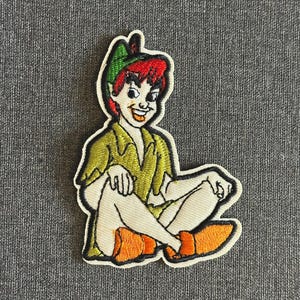 May include: Embroidered patch of a cartoon character with red hair, a green hat, and a green outfit. The character is seated with legs crossed and wearing orange shoes. The patch has a white border, suitable for clothing or accessories.