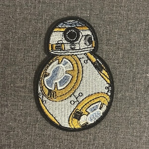 Könnte beinhalten: Gestickter Aufnäher mit dem BB-8-Droiden aus Star Wars. Der Aufnäher hat einen weißen Körper mit gelben und blauen Akzenten und einen schwarzen Rand. Das Design umfasst detaillierte mechanische Elemente.