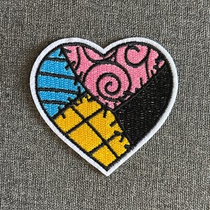 Peut inclure: Un écusson en forme de cœur avec une bordure blanche, présentant un motif patchwork. Le motif comprend des sections de rayures bleues, de tourbillons roses, de tissu noir et de carrés jaunes. L'écusson est probablement destiné aux vêtements ou aux accessoires.