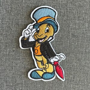 Könnte beinhalten: Gestickter Aufnäher mit einer Cartoon-Grille in Zylinder und schwarzem Jackett, die mit einer weiß behandschuhten Hand salutiert. Die Grille hat einen gelben Körper, einen blau-orangenen Hut und eine rote Krawatte. Der Aufnäher befindet sich auf grauem Stoff.