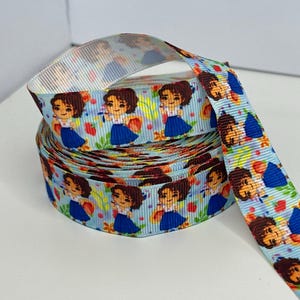 May include: A spool of light blue ribbon with a repeating pattern of a cartoon girl in a blue dress. The ribbon also features red strawberries and green leaves. The ribbon is likely used for crafts, gift wrapping, or hair accessories.