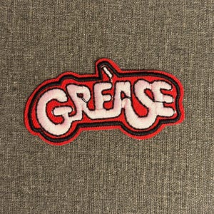 Pode incluir: Patch bordado com a palavra "GREASE" em branco, contornada em preto, sobre um fundo vermelho. O patch tem uma estética retro, ideal para jaquetas, bolsas ou outros artigos de tecido.