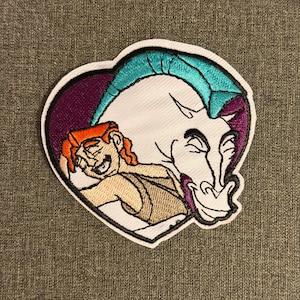 Könnte beinhalten: Gestickter Aufnäher mit Herkules und Pegasus aus dem Disney-Film. Der Aufnäher ist herzförmig mit weißem Rand. Herkules hat orangefarbenes Haar und umarmt Pegasus, der einen weißen Körper und türkisfarbene Hörner hat.