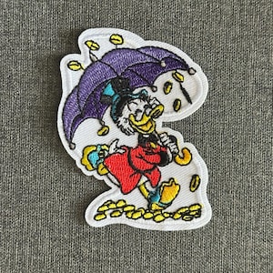 Può includere: Toppa ricamata raffigurante un'anatra dei cartoni animati con un cappotto rosso e un cappello blu, che tiene un ombrello viola. Monete d'oro e dettagli gialli circondano l'anatra. La toppa ha un bordo bianco.