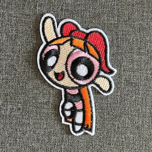 Op de afbeelding: Geborduurde patch met Blossom van The Powerpuff Girls. De patch heeft een witte rand en toont Blossom met haar rode strik, oranje haar en roze en zwarte outfit. De patch staat op een grijze stoffen achtergrond.