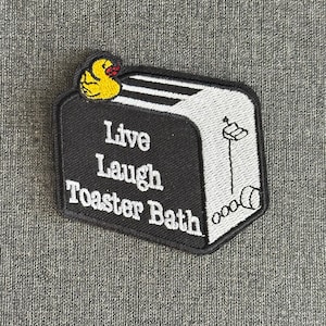 Puede incluir: Parche bordado con un diseño de tostadora en blanco y negro y las palabras "Live Laugh Toaster Bath". Un pato amarillo con un lazo rojo está encima. El parche es rectangular con un borde negro.