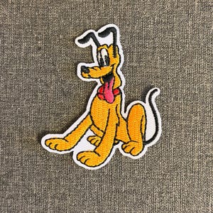 Op de afbeelding: Geborduurde patch met Pluto, een gele cartoonhond met een rode tong en zwarte oren. De patch heeft een witte rand en is geplaatst tegen een grijze stoffen achtergrond. De hond zit.