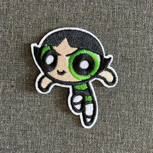 May include: Embroidered patch of Buttercup from The Powerpuff Girls. The patch has a black outline, black hair, a green eye, and a green and black outfit. The patch is on a grey fabric background.