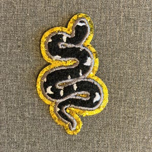 Può includere: Toppa ricamata a forma di serpente nero con segni bianchi e bordo dorato con paillettes. Il serpente ha un design curvo e stilizzato. La toppa è su uno sfondo grigio strutturato.
