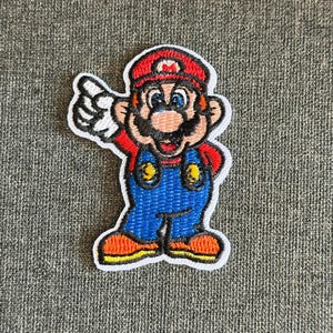 May include: Embroidered patch featuring the character Mario from the Super Mario Bros. video game series. Mario is depicted in his classic outfit: a red hat, blue overalls, and a red shirt. The patch has a white border and is on a gray background.