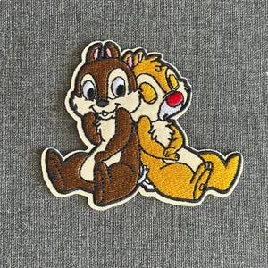 Könnte beinhalten: Gestickter Aufnäher mit Chip und Chap, den Disney-Chipmunks. Chip ist braun, Dale gelb mit roter Nase. Beide umarmen sich, mit cremefarbenem Rand. Auf grauem Hintergrund.