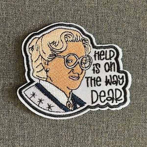 Può includere: Toppa ricamata raffigurante una donna con occhiali e uno chignon biondo. La toppa include il testo "Help is on the way Dear" in un design in bianco e nero. La toppa è bordata di nero.