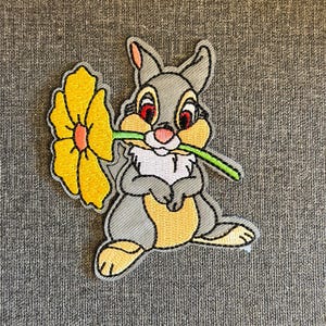 May include: Embroidered patch featuring a cartoon rabbit holding a large yellow flower in its mouth. The rabbit is gray, with a white chest and yellow accents. The flower has a yellow petal design with an orange center.