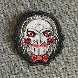 May include: Embroidered patch featuring a stylized face with red eyes, rosy cheeks, and a red bow tie. The face has white hair and a black border. The patch is likely for clothing or accessories.