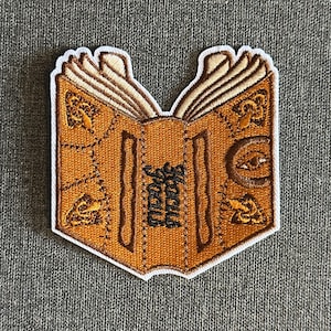 Pode incluir: Patch bordado com um design de livro aberto com a palavra "Hocus Pocus" em escrita preta. O livro é castanho com costuras castanhas e detalhes dourados, e uma borda branca. Para roupas ou acessórios.