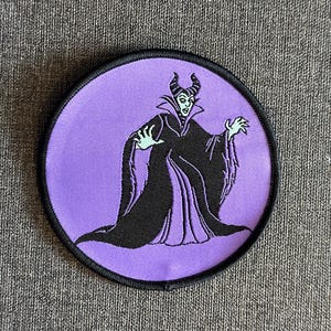 Könnte beinhalten: Ein runder Aufnäher mit einer schwarzen und violetten Illustration einer Figur mit Hörnern und einem schwarzen Umhang. Die Figur befindet sich auf einem lila Hintergrund mit schwarzem Rand. Für Kleidung oder Accessoires.