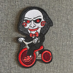 May include: Embroidered patch featuring a character from the horror film franchise Saw. The patch depicts the character riding a red tricycle. The character has a white face, red eyes, and a red bow tie, with a black cloak and hood.