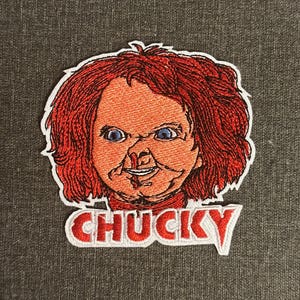 Può includere: Toppa ricamata raffigurante il volto di Chucky, con capelli rossi, occhi blu e incarnato arancione cucito. La parola "CHUCKY" è ricamata in lettere rosse con un contorno bianco. La toppa è su uno sfondo di tessuto grigio.