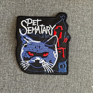 May include: Black embroidered patch with a blue cat and red eyes, and the words "Pet Sematary" in white. The patch has a distinctive shape, suitable for clothing or accessories.
