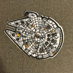 Pode incluir: Patch bordado com a nave espacial Millennium Falcon de Star Wars. O patch é branco com detalhes em preto e detalhes em laranja. O patch está em um fundo cinza texturizado.