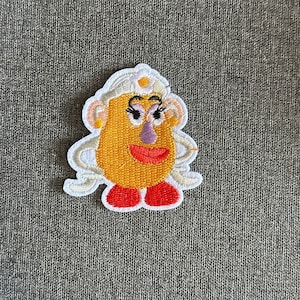 Peut inclure: Écusson brodé représentant un personnage de dessin animé avec un visage jaune, un nez violet et des lèvres rouges. Le personnage a des cheveux blancs et des pieds rouges. Le patch est bordé de blanc, pour vêtements ou accessoires.