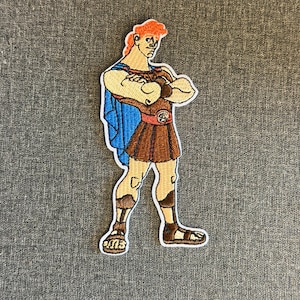May include: Embroidered patch depicting Hercules, a cartoon character. The patch shows Hercules with orange hair, a blue cape, and brown clothing. The patch is outlined in white and is on a grey background.