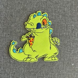 May include: Embroidered patch featuring a lime green cartoon dinosaur holding a slice of pizza. The dinosaur has an orange eye, blue nose, and blue accents. The pizza slice has red, yellow, and brown toppings. The patch is outlined in black.