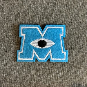 May include: A light blue felt patch in the shape of the letter "M" with a white border. The "M" features a white-outlined eye with a black pupil. The patch is likely for a Monsters, Inc. theme.