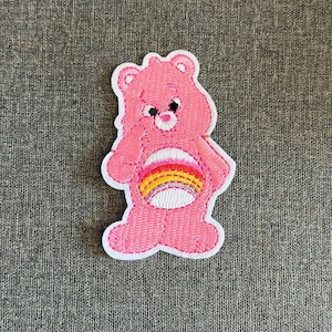 May include: A pink embroidered patch featuring a cartoon bear with a rainbow belly. The bear has a white outline and is set against a gray textured background. The bear is in a standing pose with one paw raised.