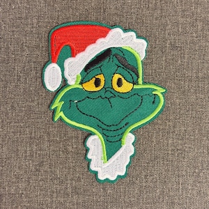 May include: Embroidered patch of a green character with a grumpy expression, wearing a red Santa hat with white trim. The character has yellow eyes and a white collar. Suitable for clothing or accessories.