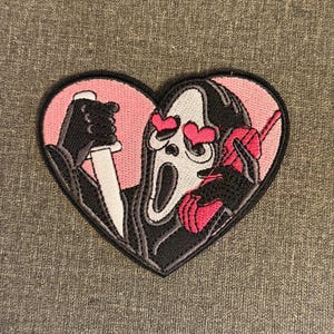 May include: Embroidered heart-shaped patch featuring the Ghostface mask from the Scream films. The mask has heart-shaped eyes and is holding a knife and a phone. The patch is black, white, and pink.