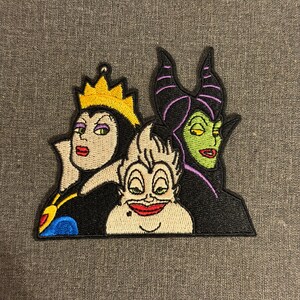 Könnte beinhalten: Gestickter Aufnäher mit drei Disney-Bösewichten: Die böse Königin, Ursula und Maleficent. Der Aufnäher hat einen schwarzen Rand und zeigt die Charaktere in leuchtenden Farben, darunter Gold, Lila, Grün und Rot. Die böse Königin trägt eine goldene Krone.