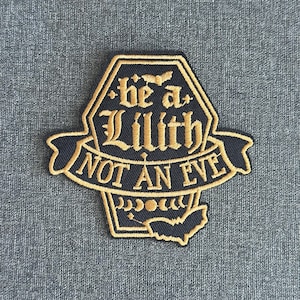 May include: Embroidered patch featuring the words "be a Lilith NOT AN EVE" in gold on a black background. The patch has a distinctive shape with a banner and decorative details.