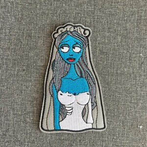 May include: Embroidered patch featuring a blue-skinned bride with gray hair and a white wedding dress. The patch has a gray border and is likely inspired by a Tim Burton film. The bride has large eyes and red lips.