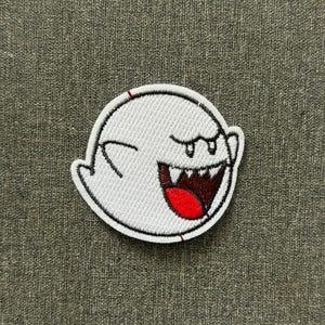 Puede incluir: Un parche bordado circular que presenta un personaje de fantasma blanco con un contorno negro, una boca muy abierta que muestra dientes afilados y una lengua de color rojo brillante. El diseño está cosido con hilo blanco denso sobre una base.