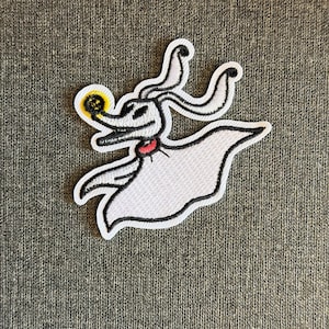 May include: Embroidered patch of Zero from The Nightmare Before Christmas. The patch shows Zero, a ghost dog with a white body, black outline, and a small red collar. He has a glowing jack-o'-lantern nose. The background is grey.